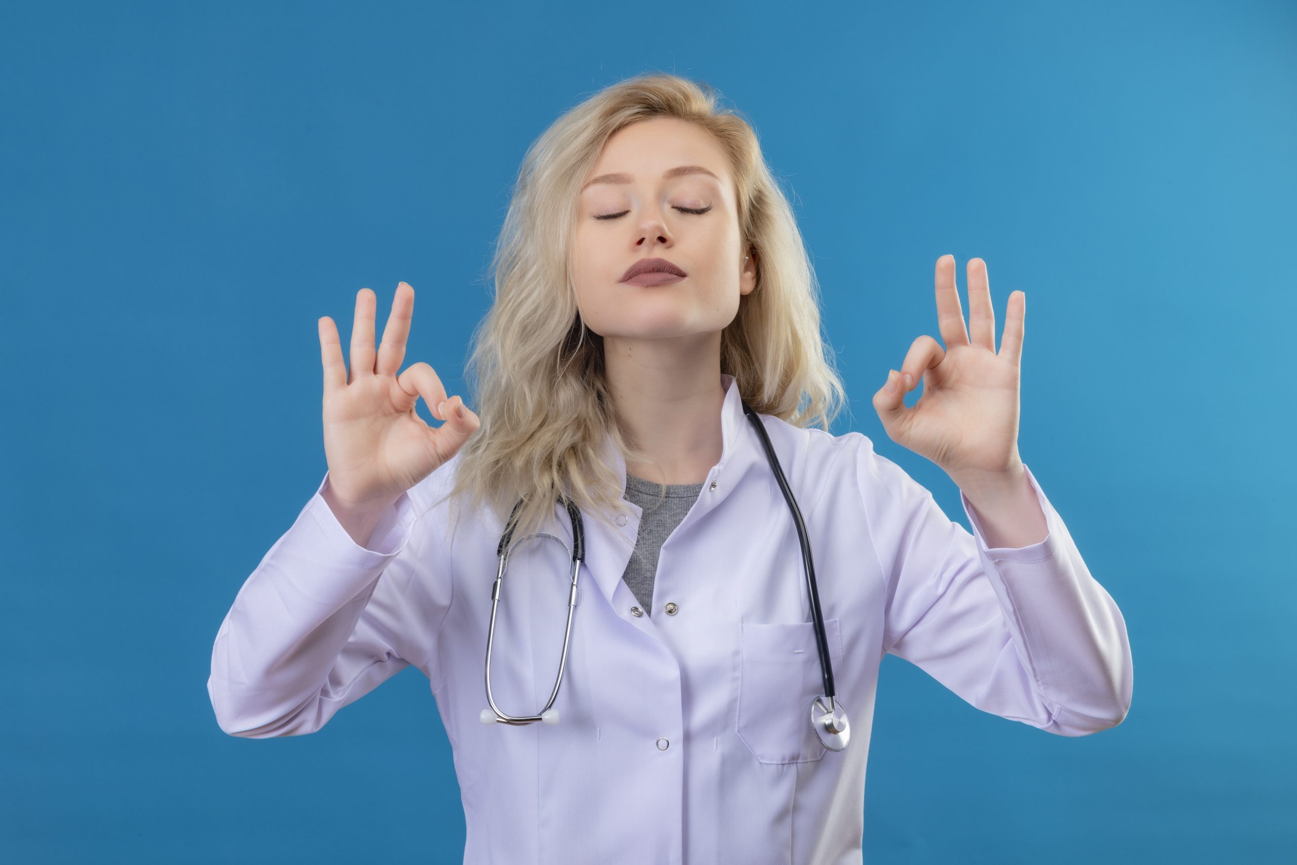 with closed eyes doctor young girl wearing stethoscope in medical gown showing okey gesture on blue background